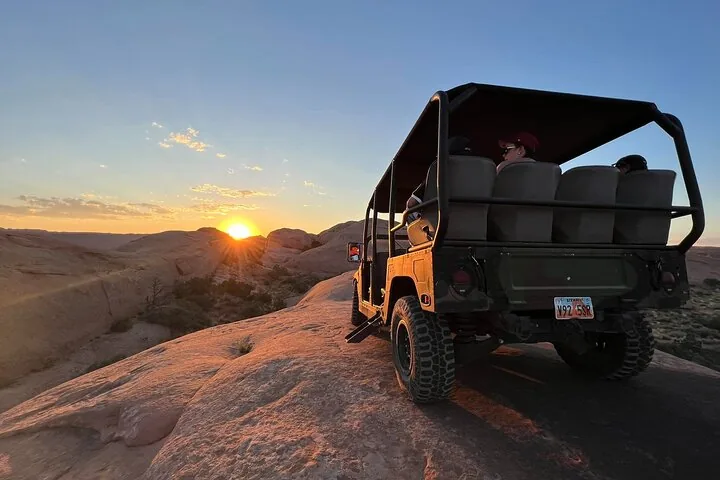 Passagiere im Heck eines Offroad-Fahrzeugs blicken über Felsen in Richtung Sonnenuntergang.